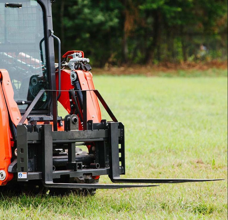 A close-up of a skid steer loader equipped with black heavy-duty pallet forks, parked in a grassy field.
