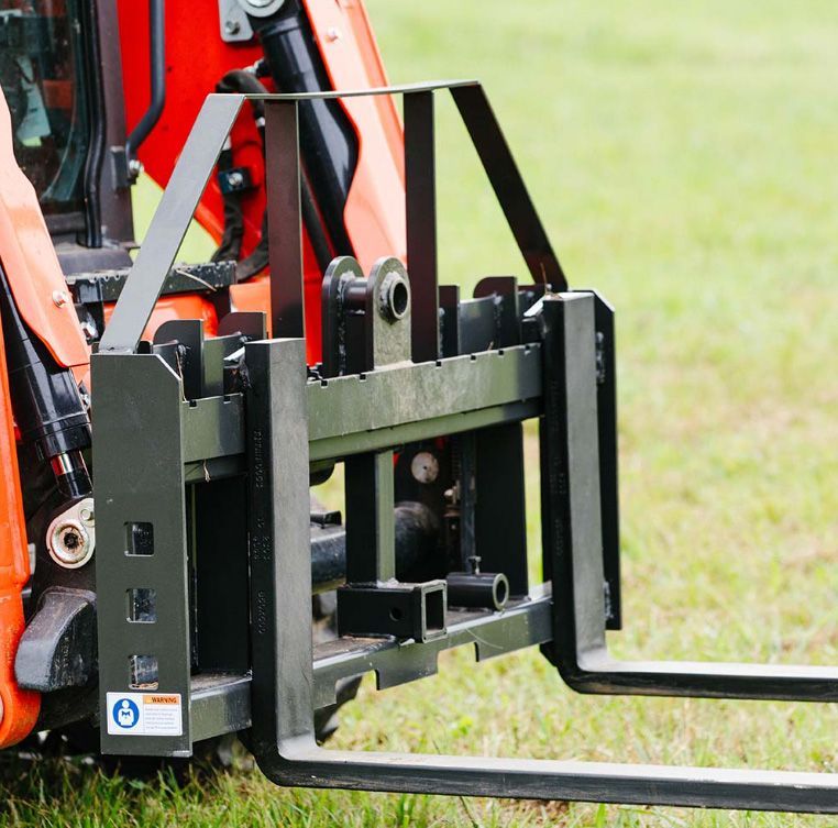 A black metal pallet fork attachment mounted to the front of an orange skid-steer loader on a grassy field.