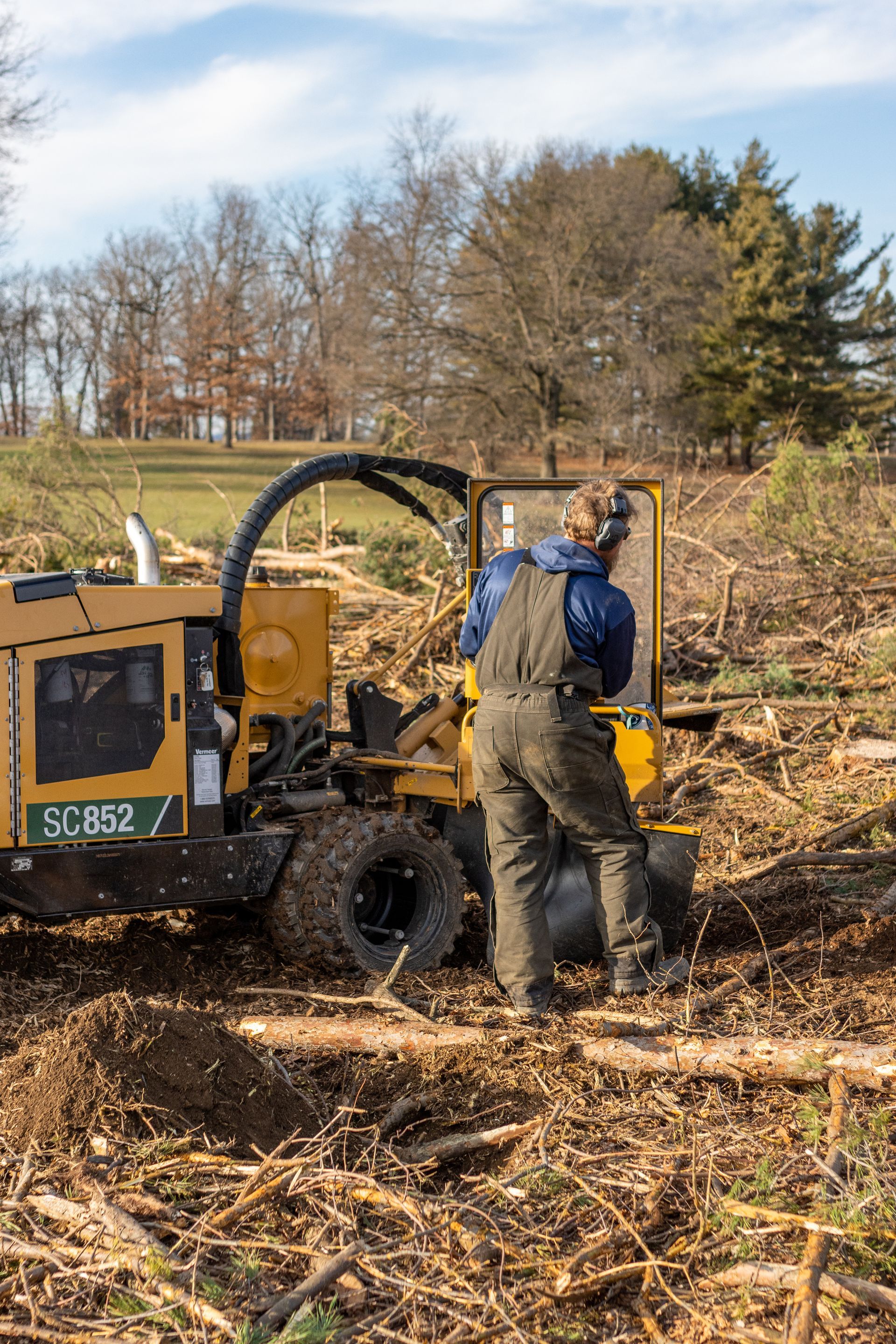 A person in brown overalls operates a yellow Vermeer stump grinder in a clearing covered in wood chips and debris.
