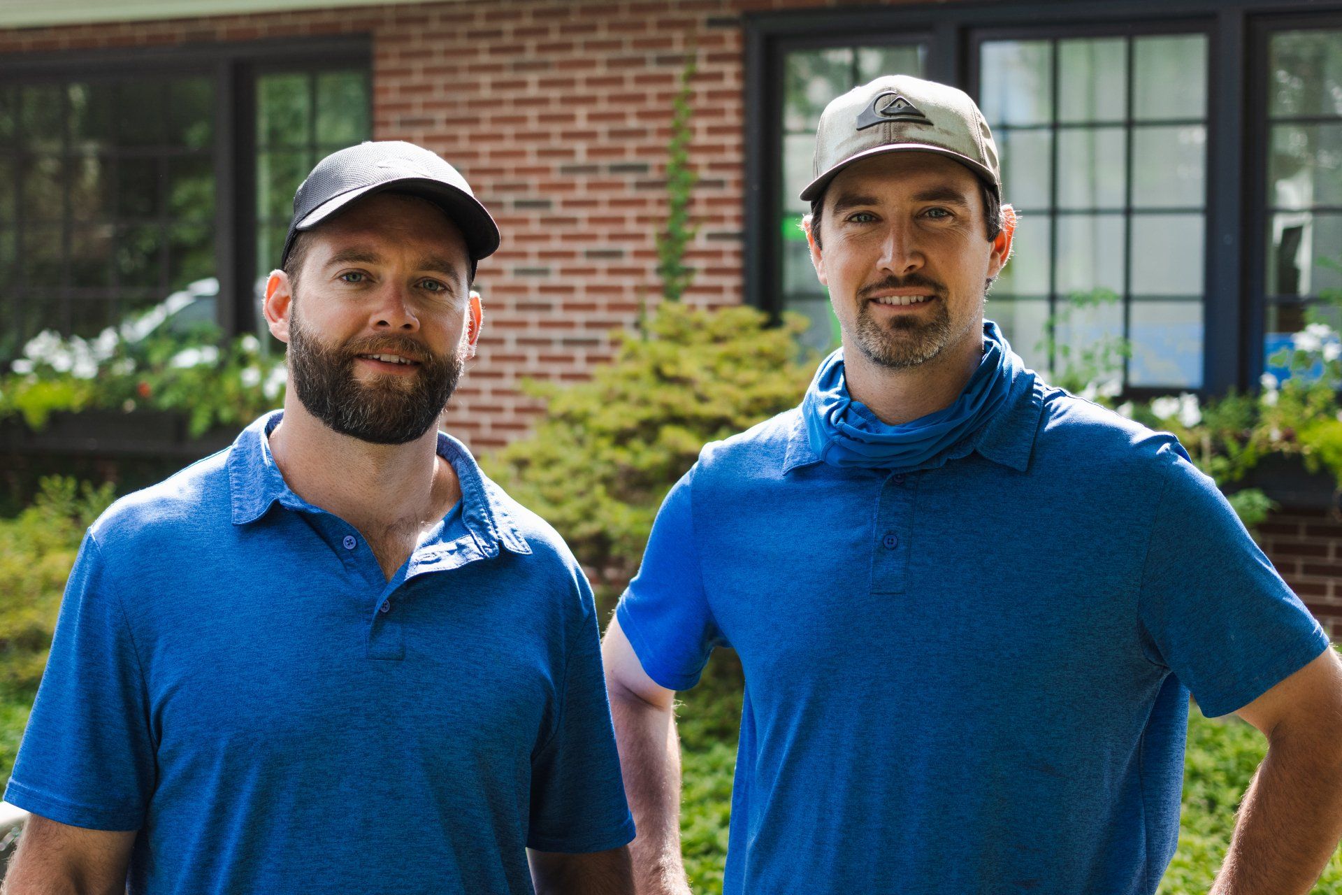 Two men in blue polo shirts and baseball caps pose together in front of a brick building and greenery.