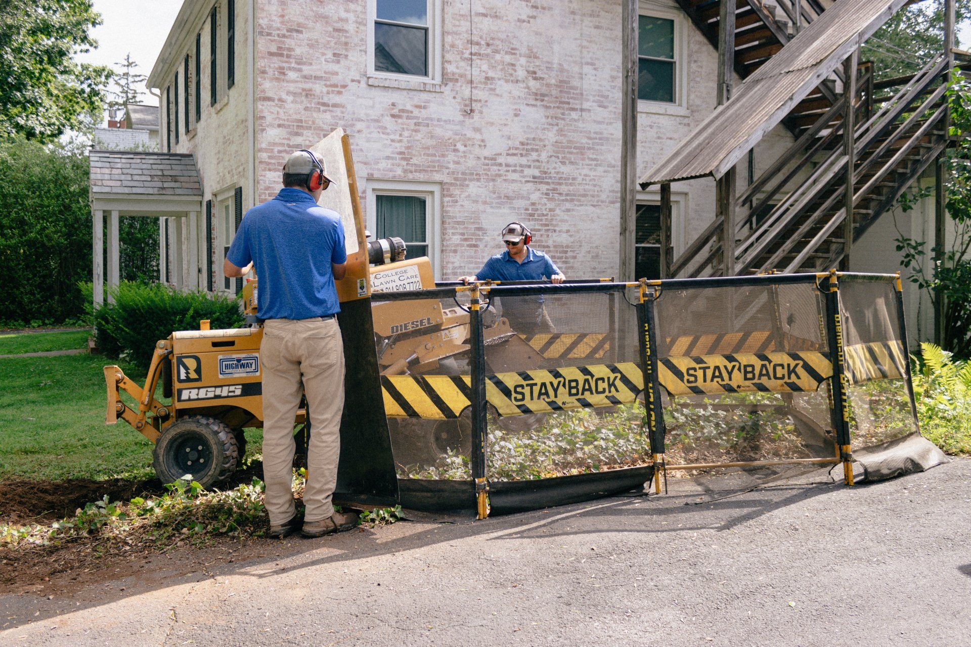 Two workers in safety gear operate a stump grinder protected by a caution screen near a stone house.