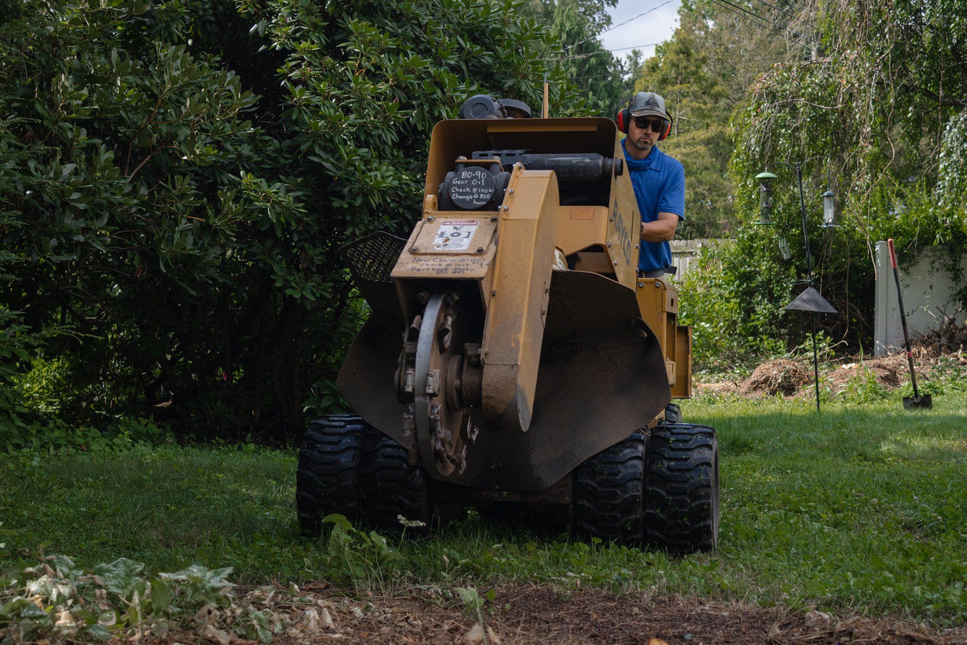 A worker in a blue shirt uses a yellow stump grinder to remove a tree stump in a grassy yard.