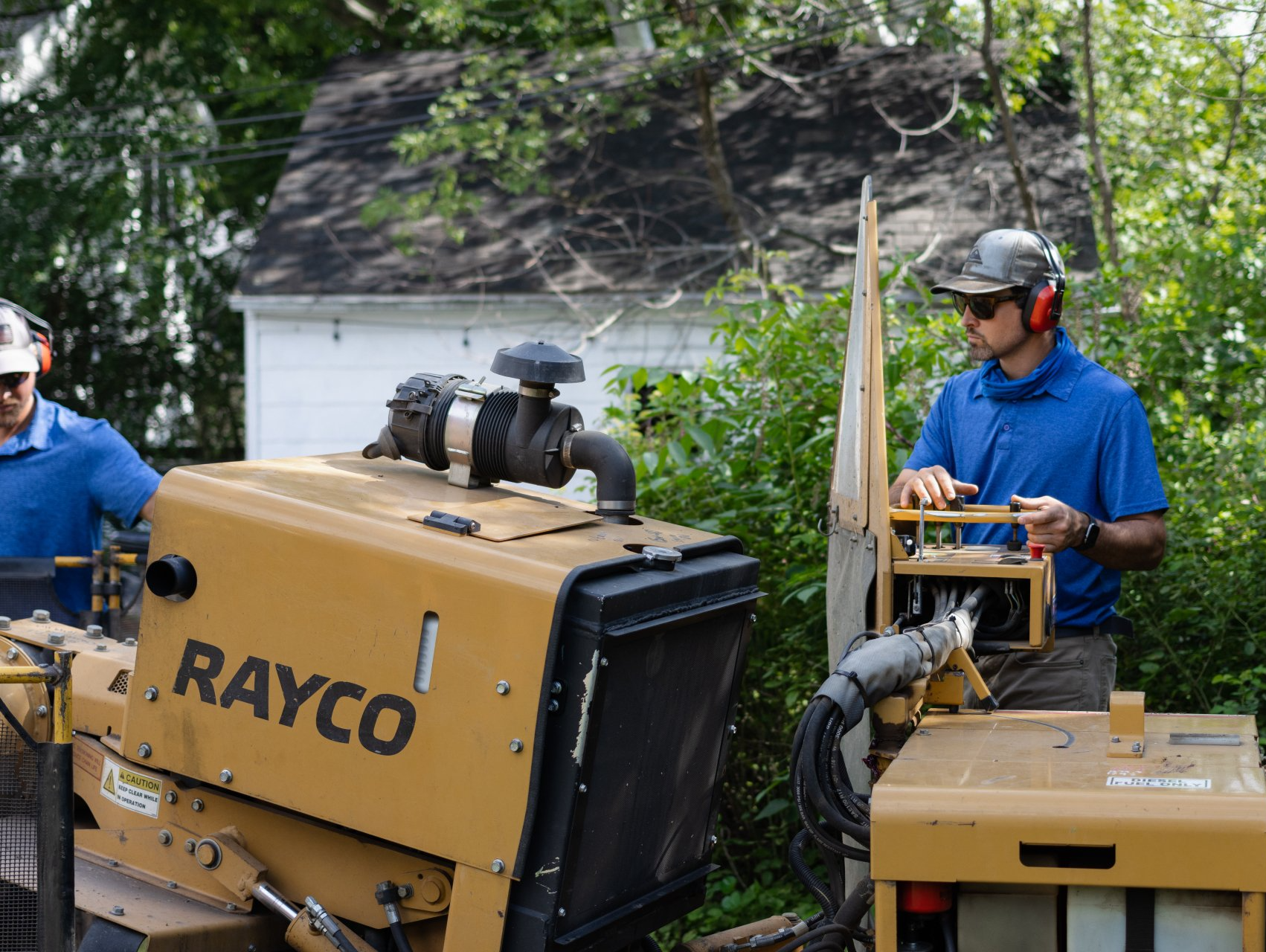 Two workers wearing ear protection operate a yellow Rayco stump grinder in a wooded residential area.