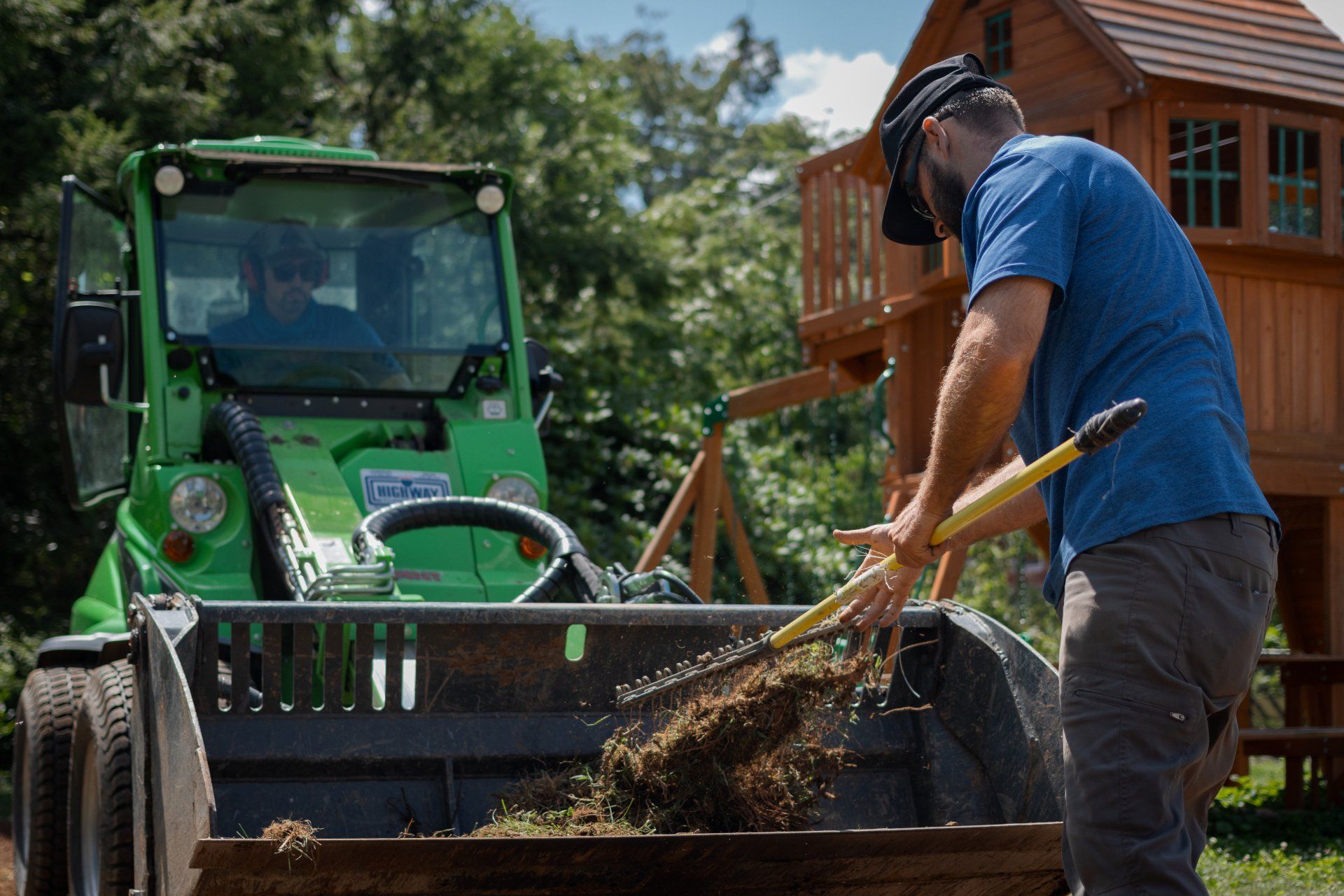 A person uses a garden rake to load dried grass and debris into the front bucket of a green compact loader outdoors.