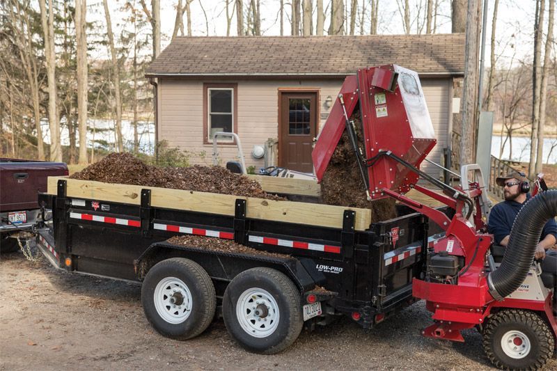 A person operates a red leaf vacuum, blowing leaves into a large black utility trailer in a wooded, outdoor setting.