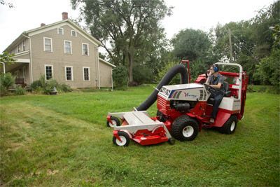 A person operating a red and white Ventrac mower with a large collection chute on a grassy lawn near an old brick house.