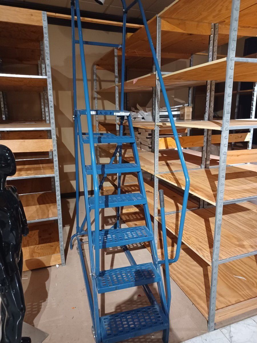 A tall, blue metal rolling ladder with handrails, positioned in an aisle between rows of wooden industrial shelving.