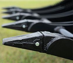 A row of black, pointed metal agricultural ripper shanks with replaceable tips aligned against a grassy field background.