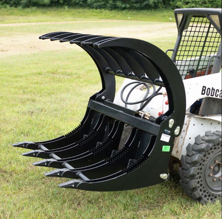 A black heavy-duty root grapple attachment mounted on a white Bobcat skid steer loader in a grassy field.