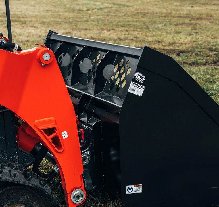 A close-up of a bright orange tractor loader arm attached to a heavy-duty black metal bucket on a grassy field.