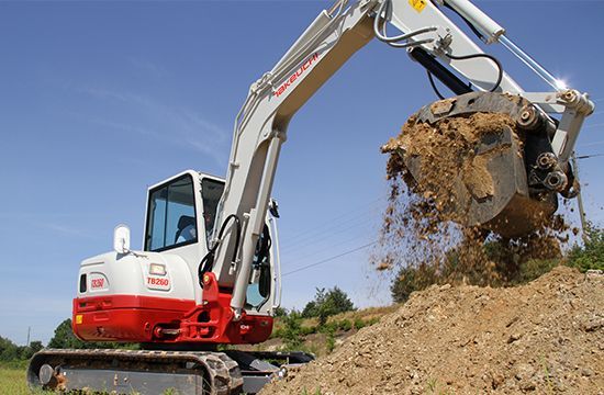 A white and red Takeuchi excavator dumps a bucket of dirt onto a pile against a clear blue sky.