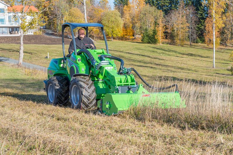 A person drives a green articulated tractor with a front-mounted flail mower across a field on a sunny autumn day.