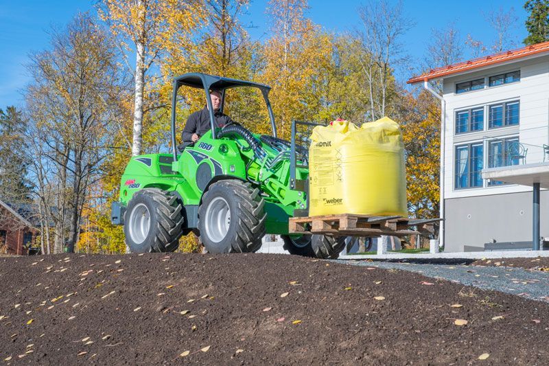 A person operating a green compact loader carrying a large yellow sack on a wooden pallet in an outdoor residential area.