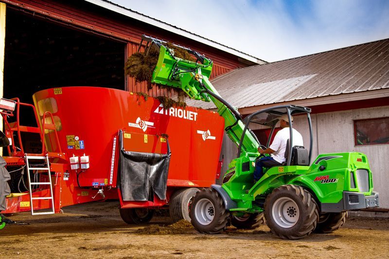 A green Avant compact loader dumping hay into a large red Trioliet TMR mixer on a farm.