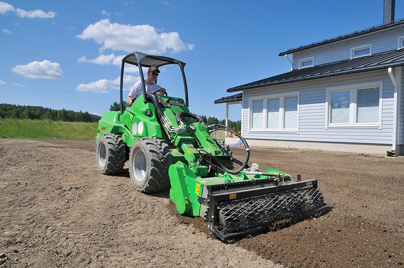 A person operates a green Avant compact loader equipped with a soil cultivator in front of a house on a sunny day.
