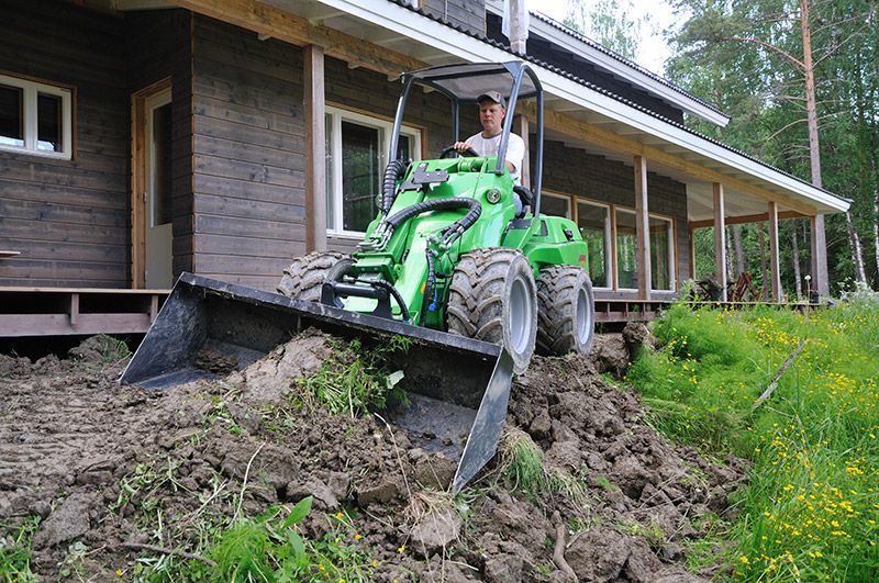 A green compact loader sits on a mound of dirt in front of a dark-paneled house with a wooden porch.