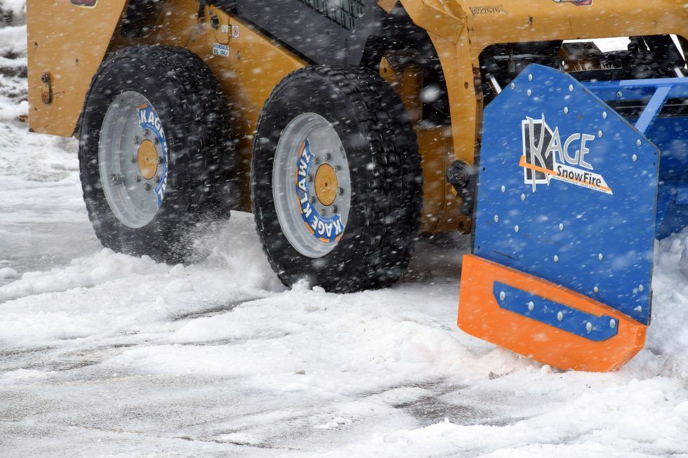 A yellow skid steer with a blue and orange Kage snow plow attachment pushing snow on a snowy surface.