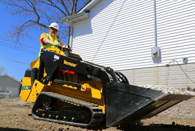 A worker in a high-visibility vest and hard hat operates a yellow Vermeer tracked loader outdoors near a white building.