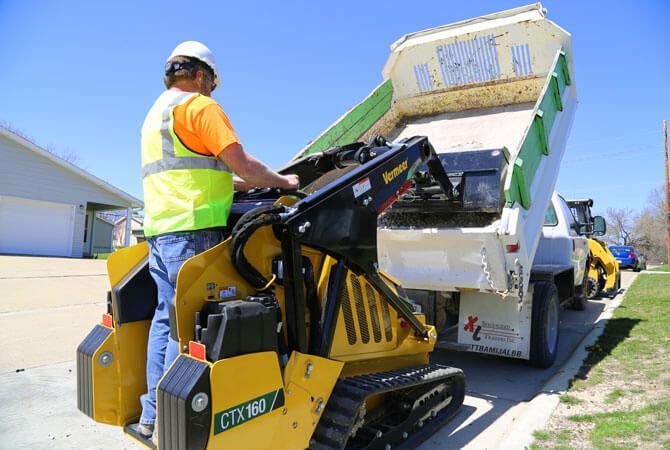 A construction worker in a high-visibility vest operates a yellow track loader, dumping materials into a truck bed.