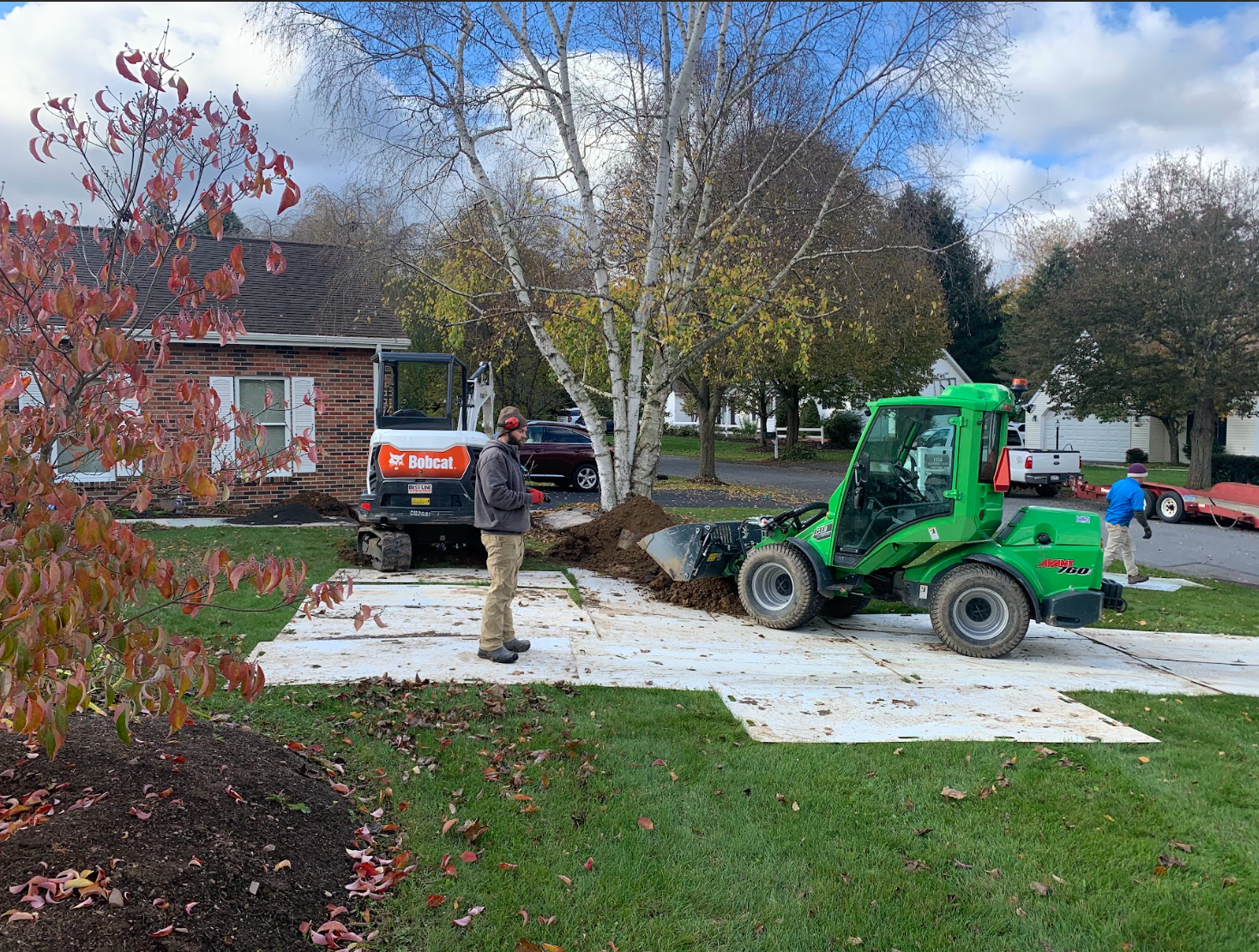 Construction workers use heavy machinery to excavate soil in a residential front yard on a sunny autumn day.