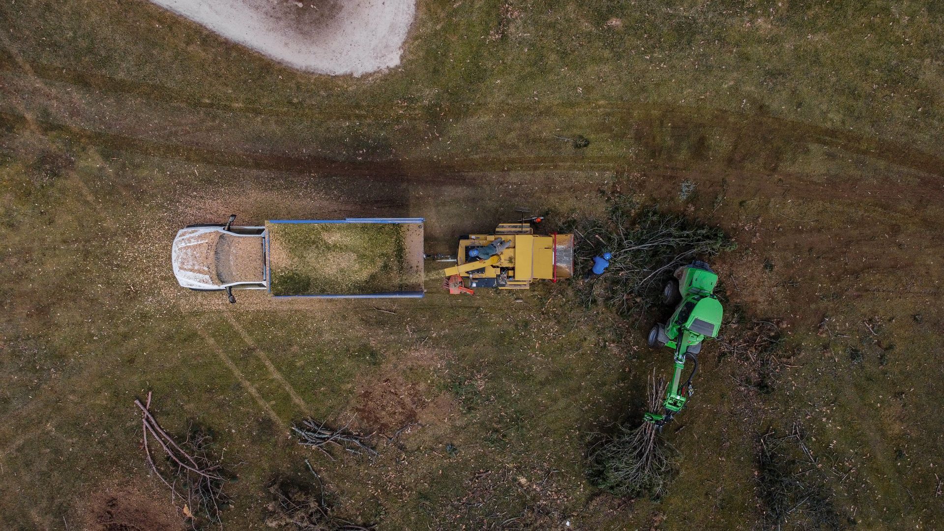 An aerial view of a green forestry tractor feeding tree branches into a yellow woodchipper, which empties into a truck.