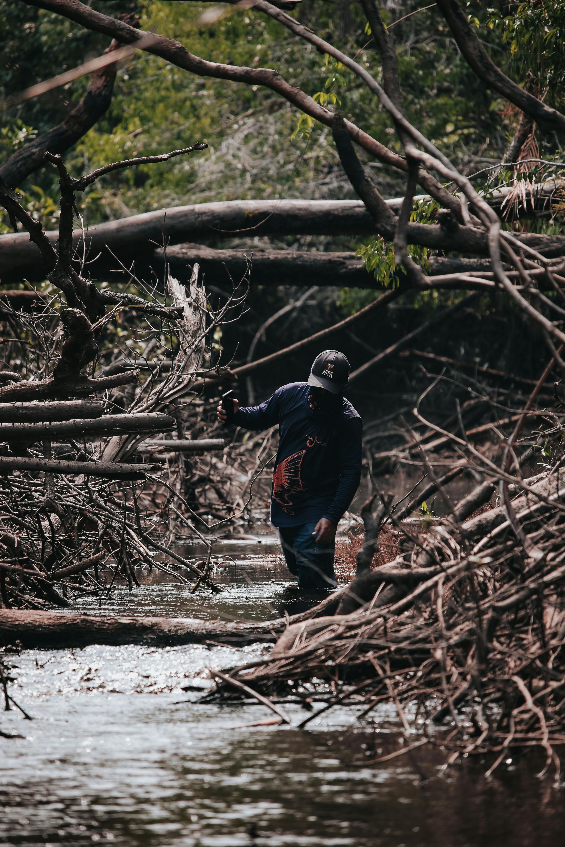 A person wearing a dark jacket and hat walks through a narrow, debris-filled forest stream surrounded by fallen branches.