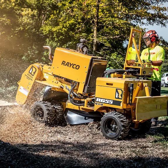 A person wearing safety gear operates a yellow Rayco RG55 stump grinder, turning a tree stump into wood chips.