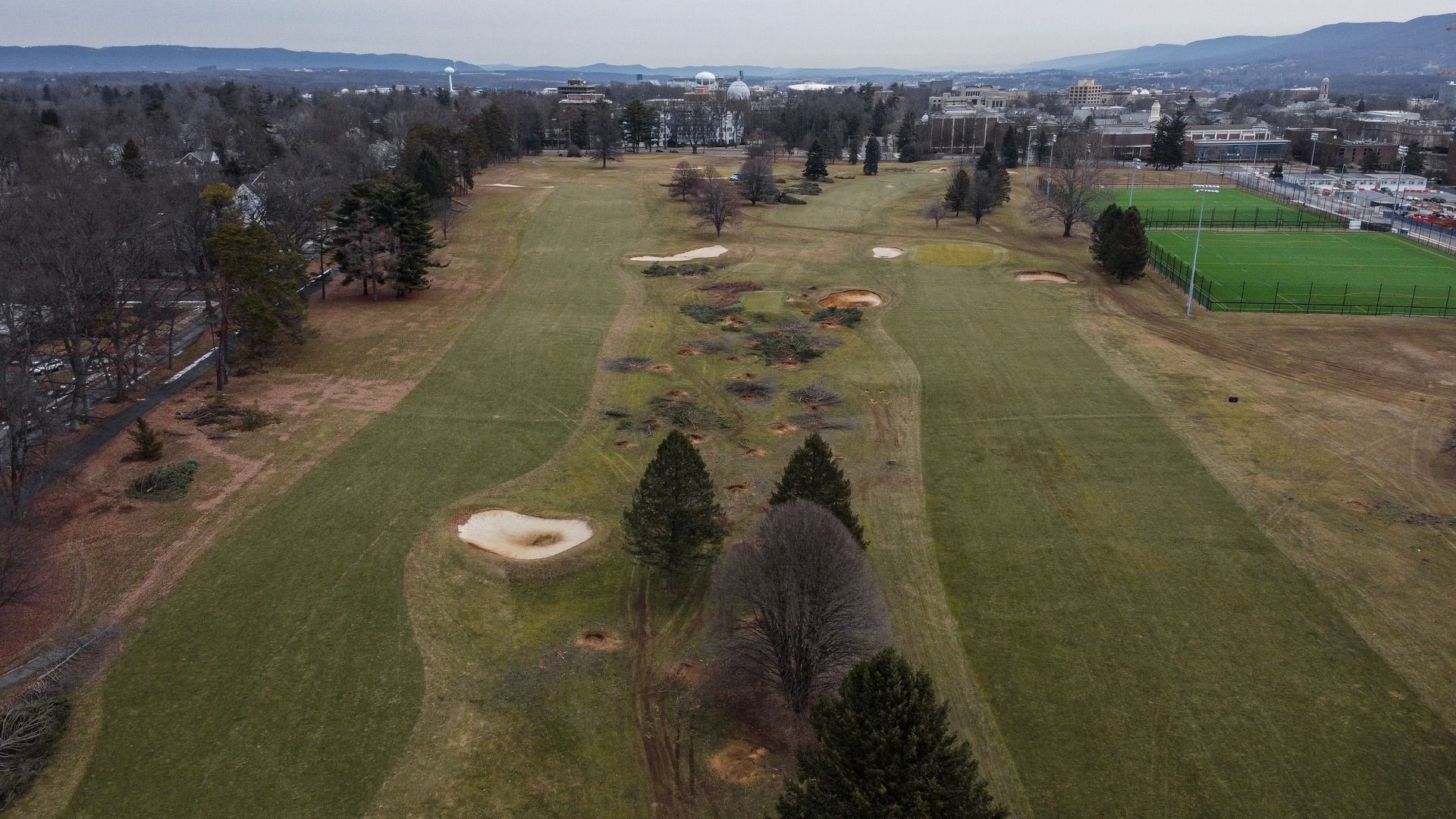 An aerial view of a golf course fairway with sand bunkers, scattered trees, and adjacent green athletic fields.
