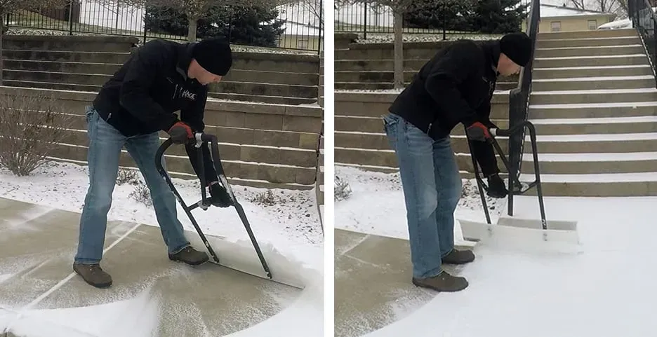 A person in winter gear uses a wheeled, long-handled snow pusher to clear snow from a paved walkway.