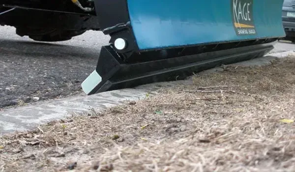 A close-up view of a blue KAGE snow plow blade edge clearing snow along a concrete curb next to a dry grassy area.