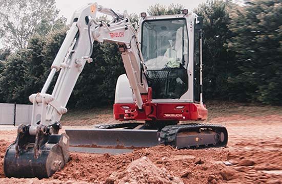 A Takeuchi mini excavator sits on a dirt construction site with a white cab, red frame, and black tracks.