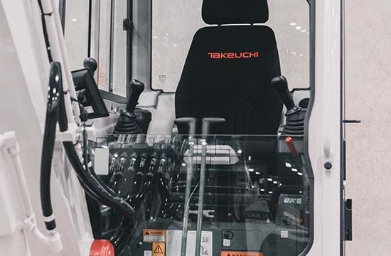 The interior cab of a Takeuchi excavator, featuring a black driver's seat, joysticks, and control panels.