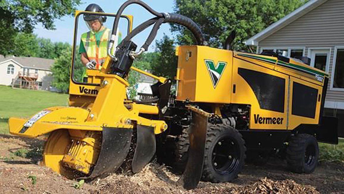 A yellow Vermeer stump grinder being operated on a grassy residential lawn near a house.