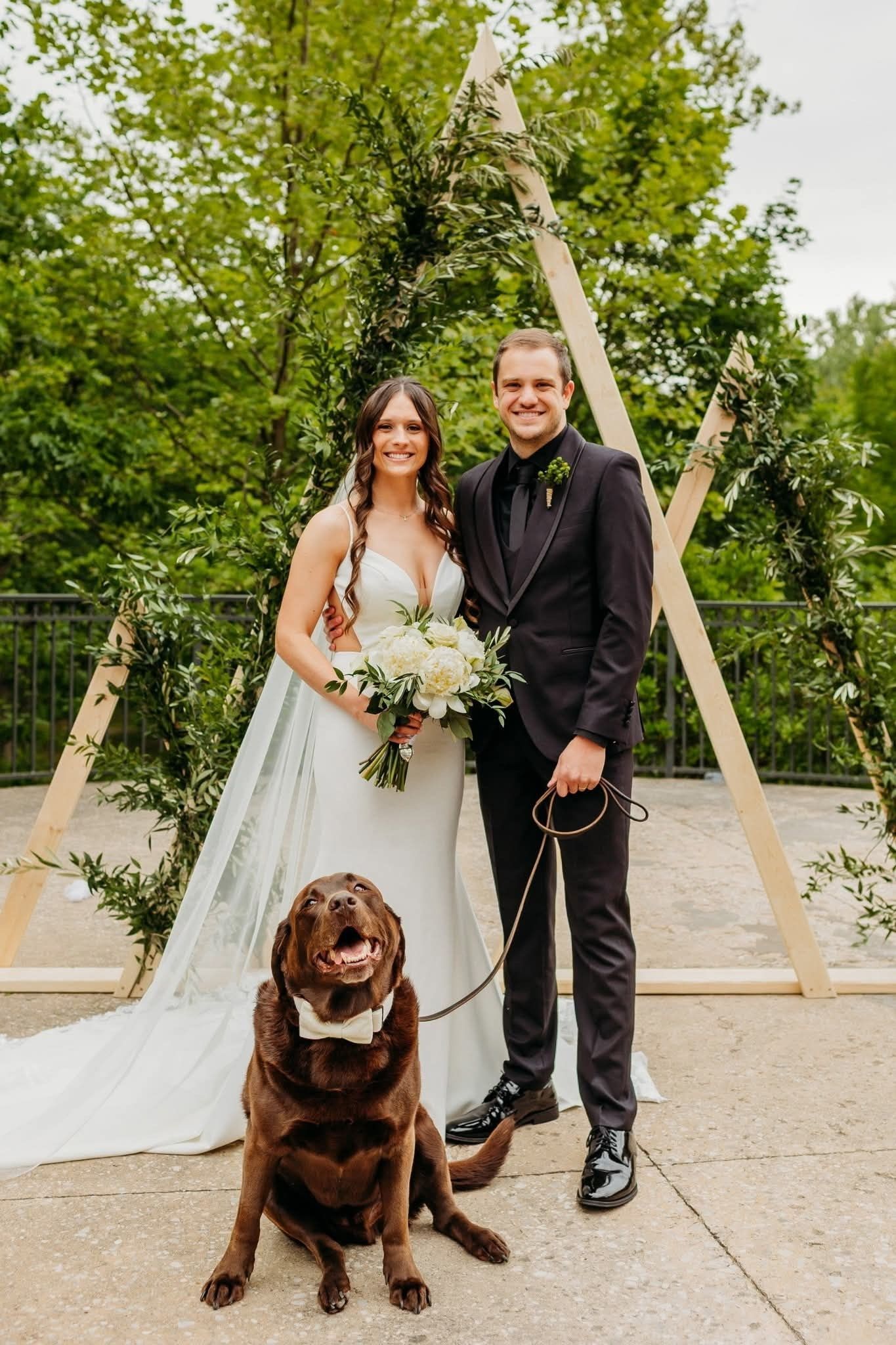A bride and groom stand outdoors with a large brown dog sitting in front of them at an altar decorated with greenery.