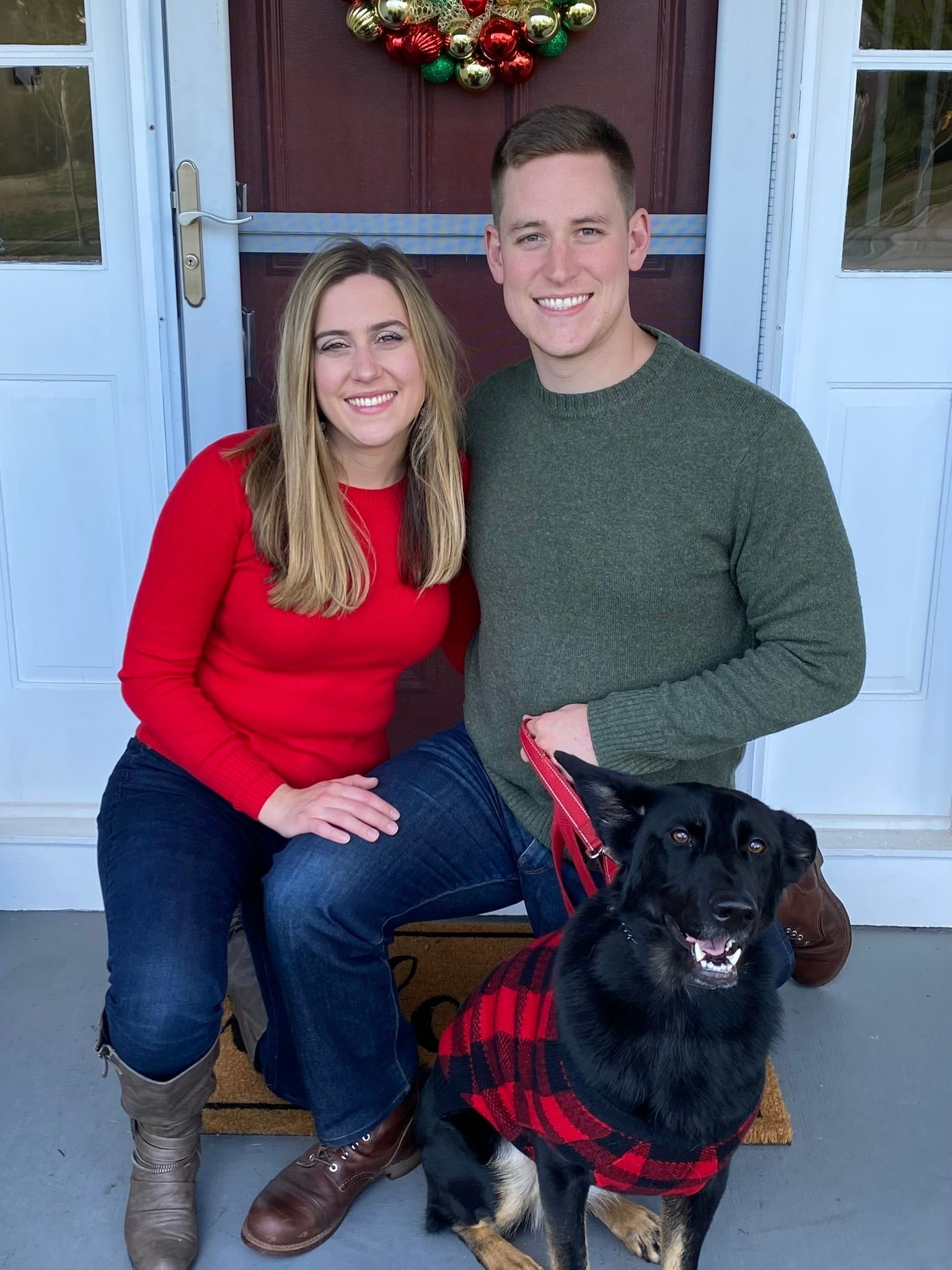 A couple in a red sweater and green sweater smile while sitting on a porch with their dog wearing a plaid coat.