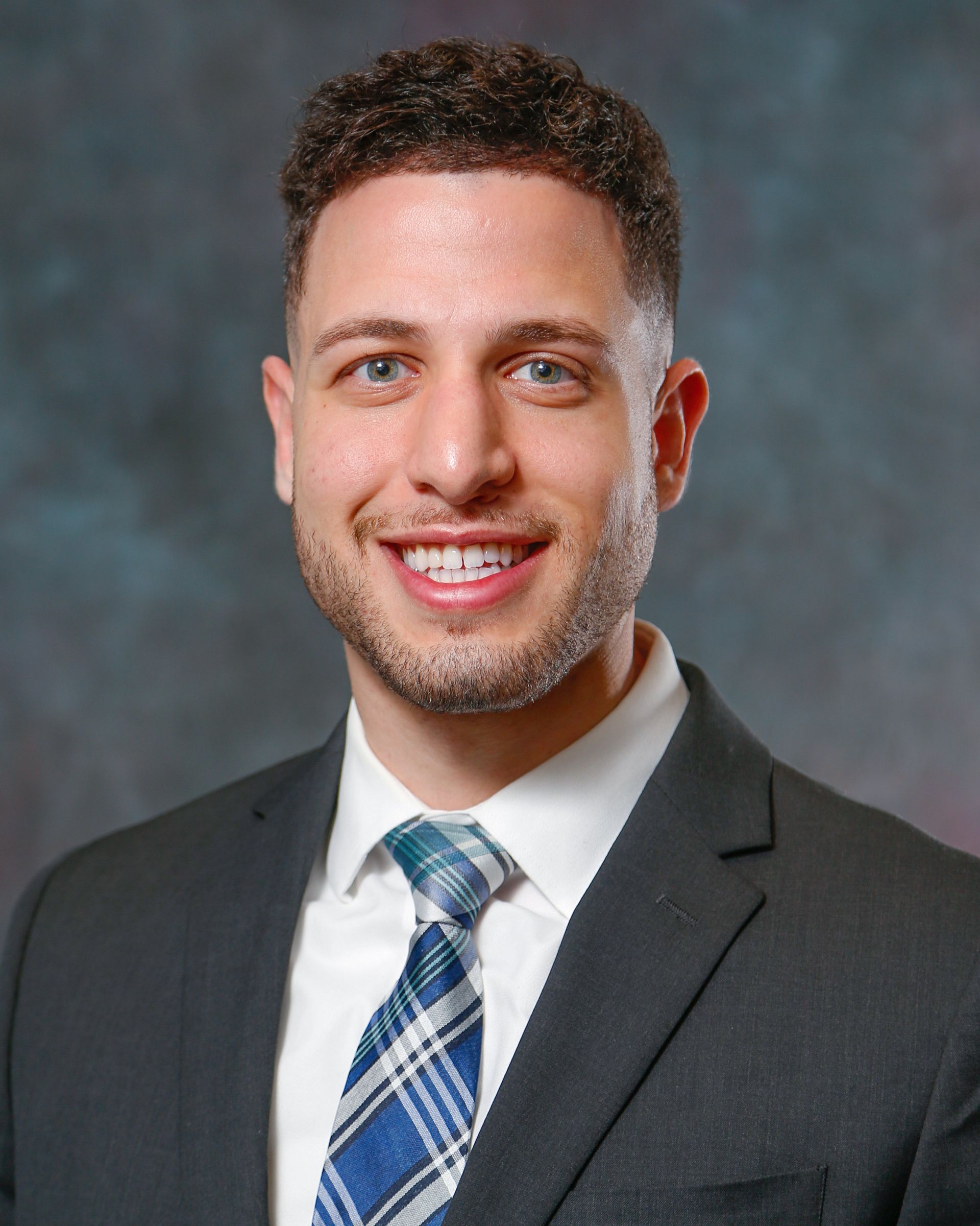 A man with short, curly hair and a beard, wearing a gray suit and plaid tie, smiling against a dark textured background.