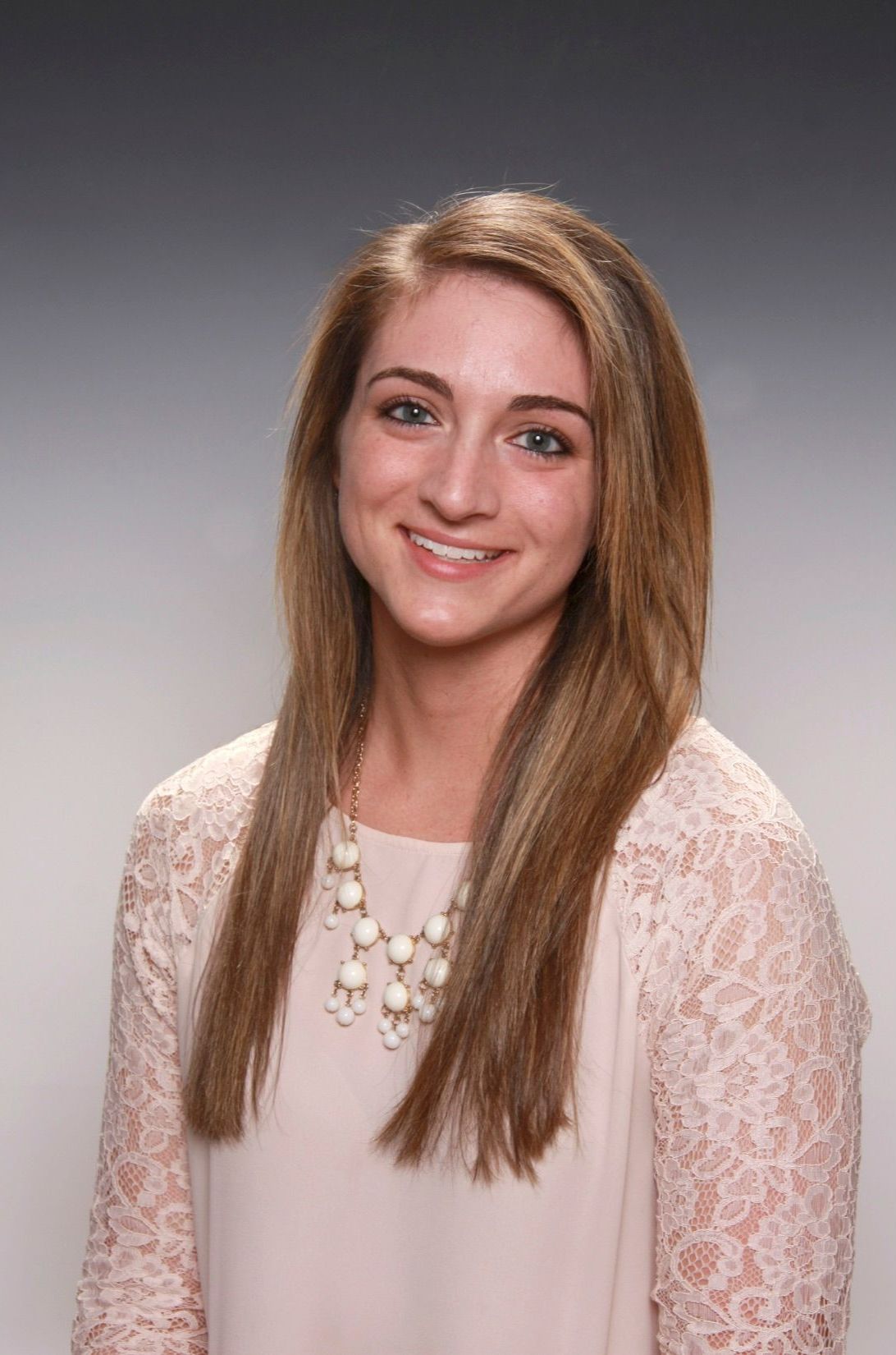 A person smiling, wearing a light pink, lace-sleeved top and a layered pearl-style necklace against a gray background.