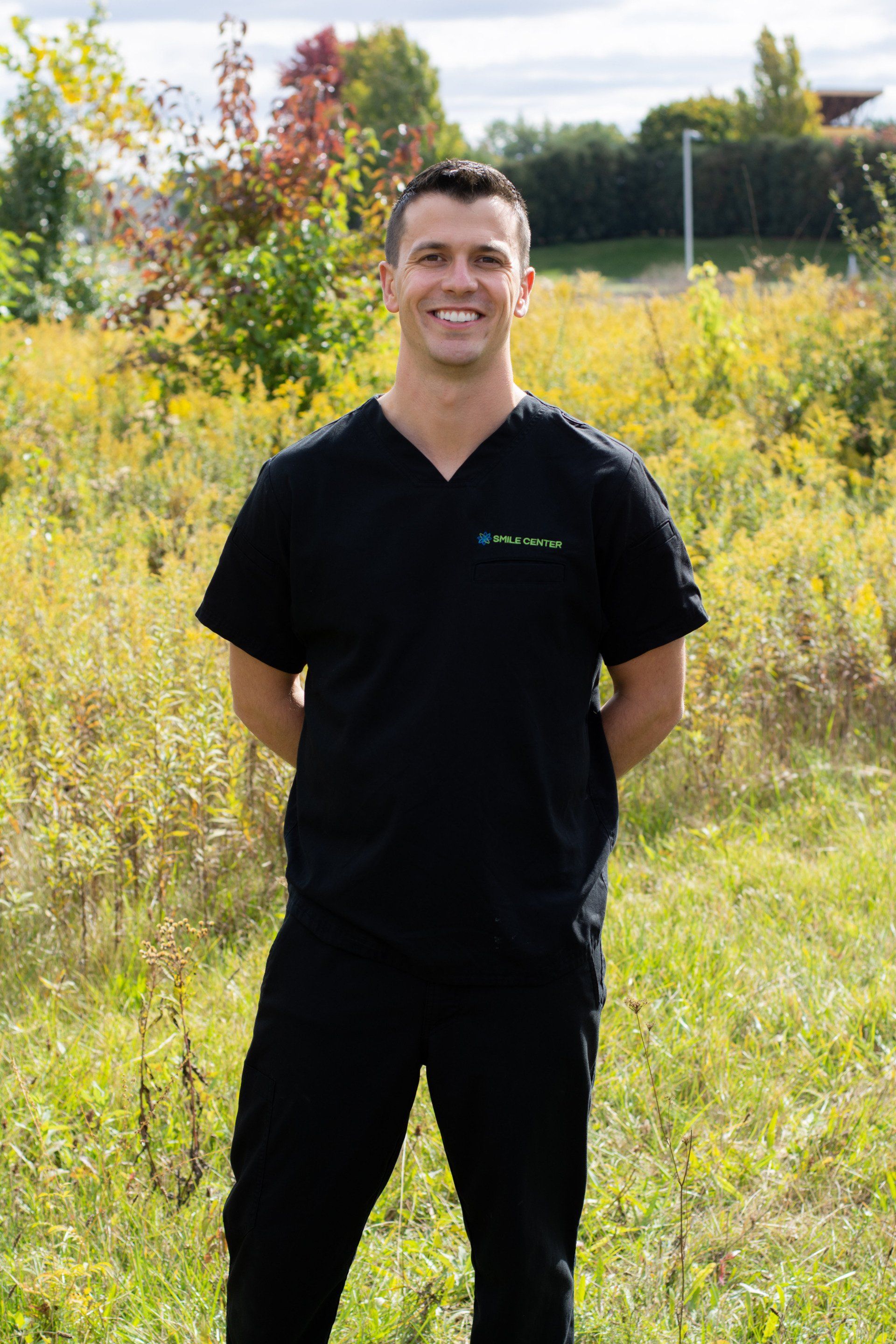 A person in black scrubs stands with arms behind their back, smiling, in a grassy field outdoors.