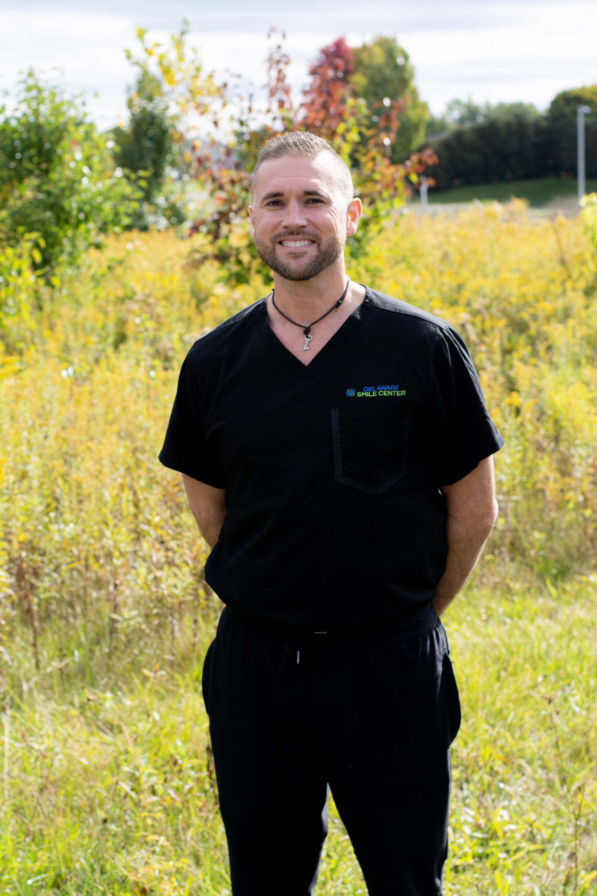 A smiling person wearing black medical scrubs stands outdoors in a field of tall, yellow-green grass under a cloudy sky.