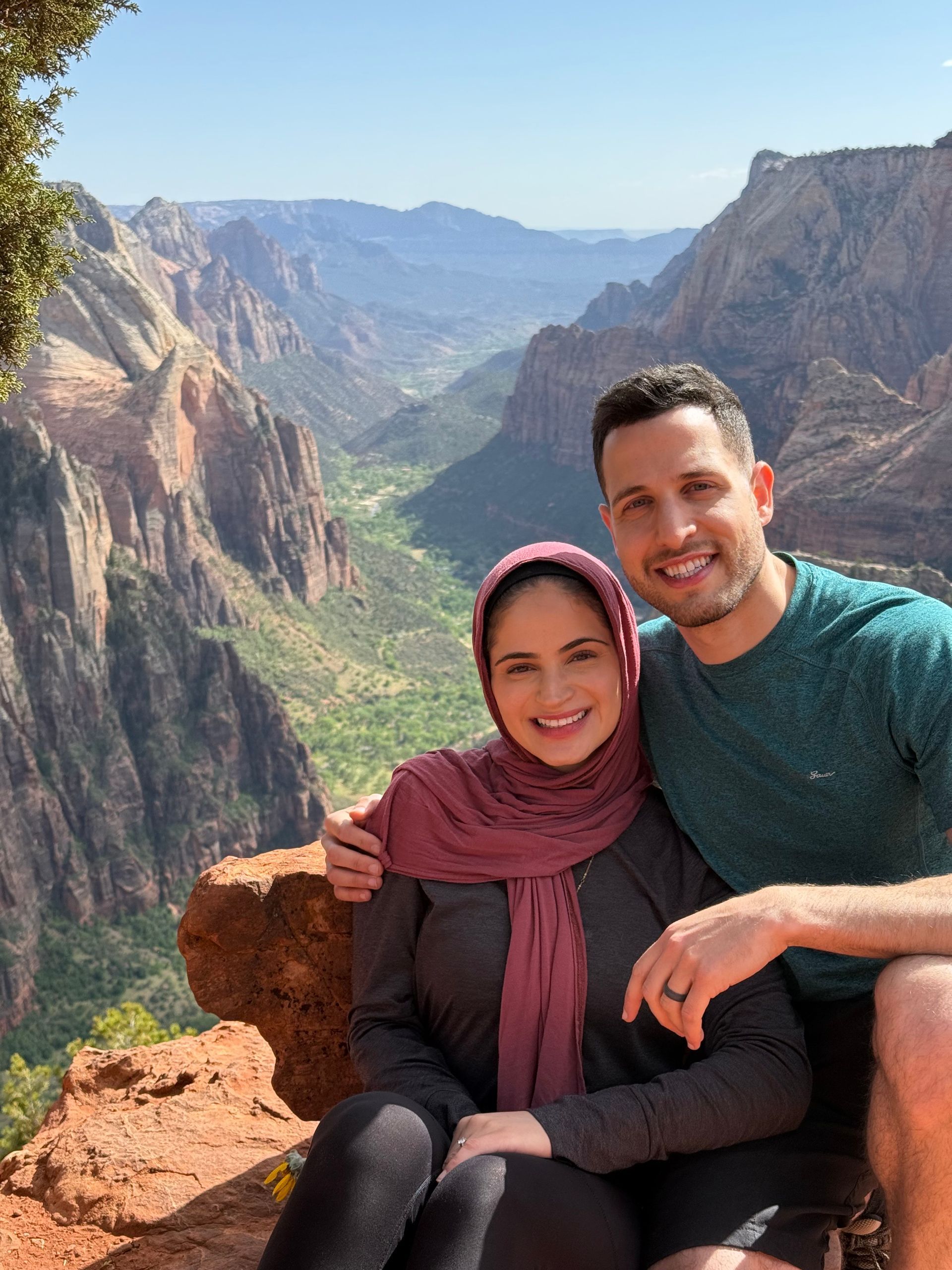A smiling couple poses for a photo on a rocky ledge overlooking a deep, expansive canyon valley under a clear sky.