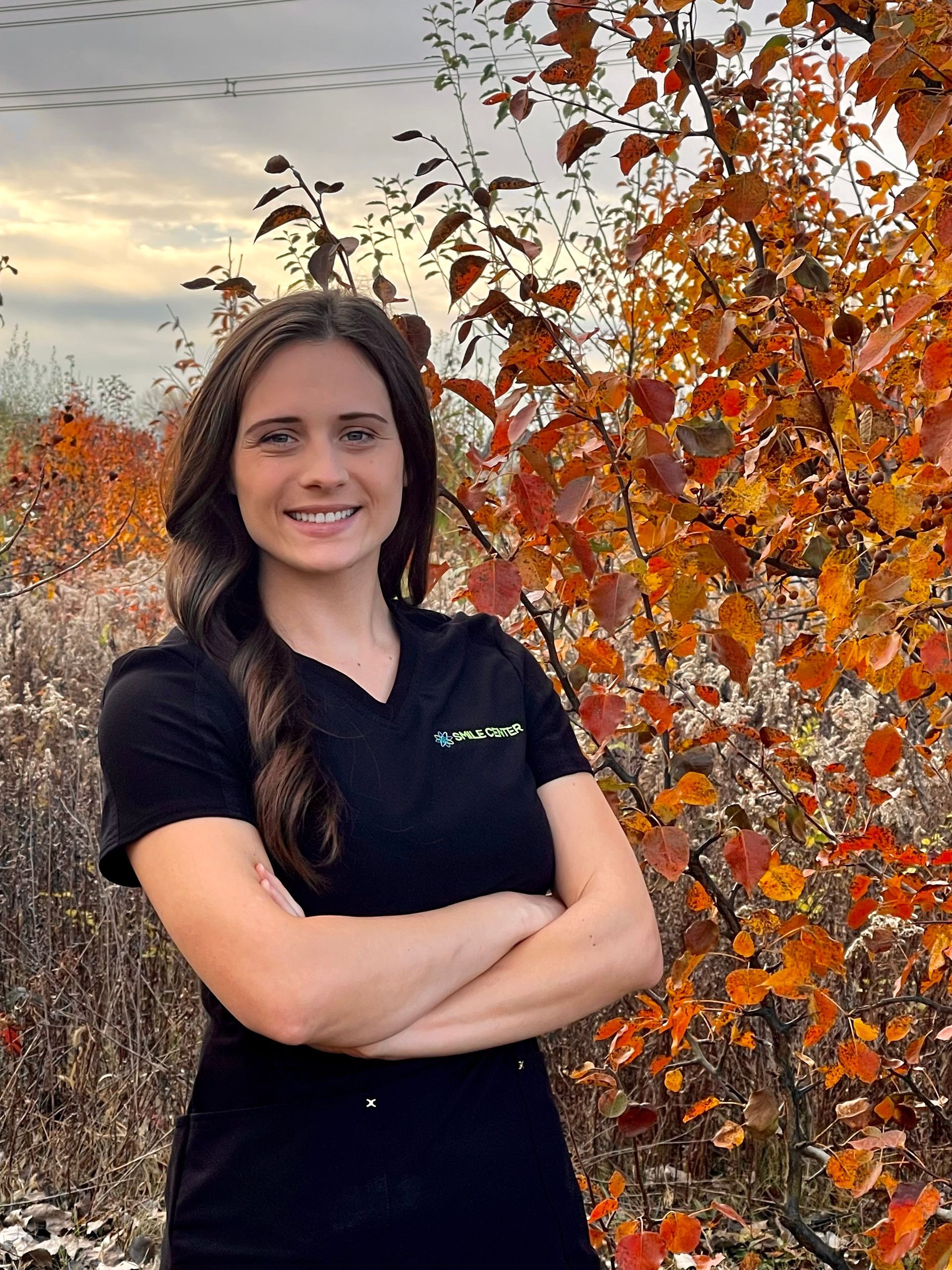 A smiling person with arms crossed, wearing a black polo shirt, stands outdoors in front of a tree with orange fall leaves.