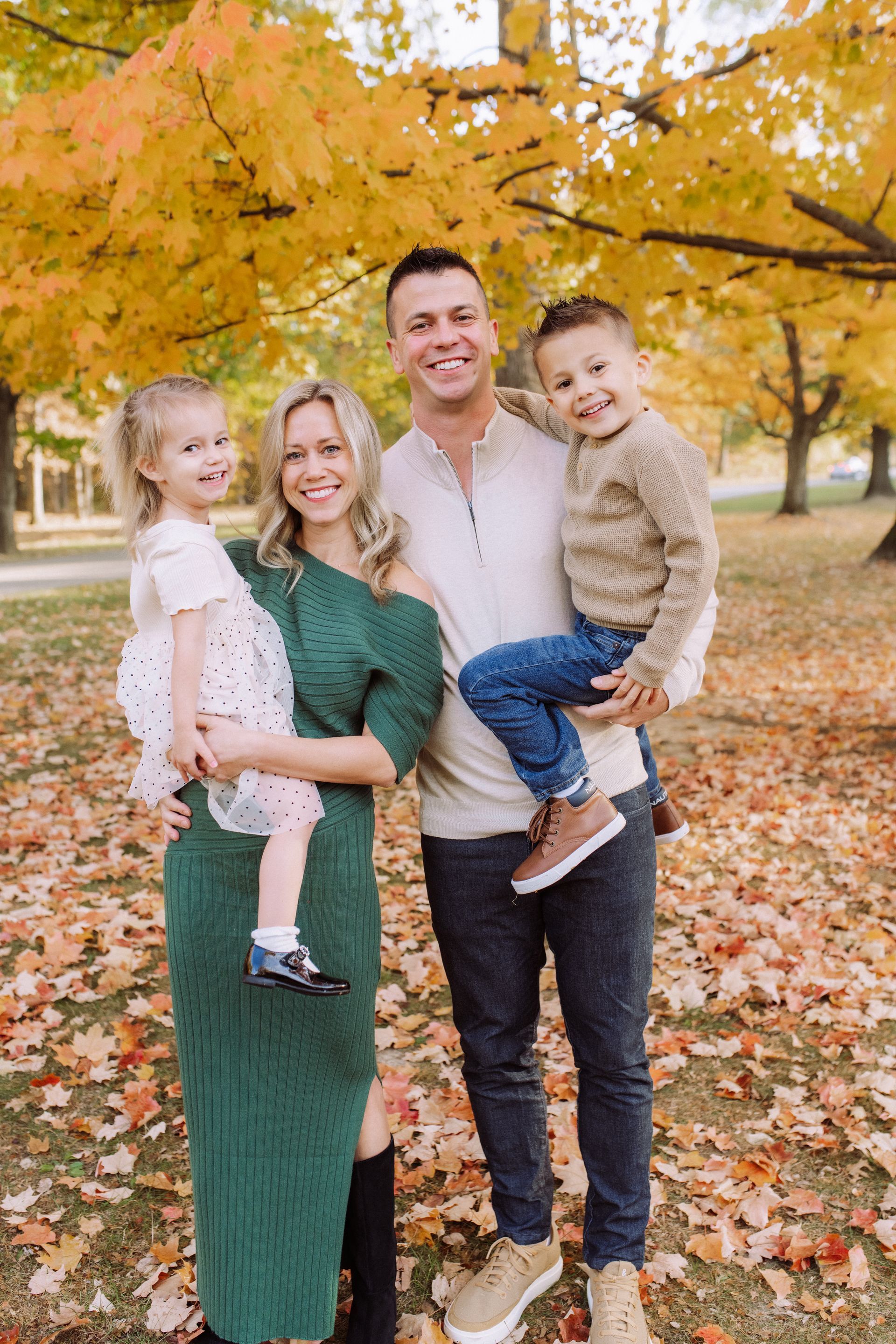 A family of four poses in a park during autumn with yellow-leafed trees and fallen leaves on the ground.