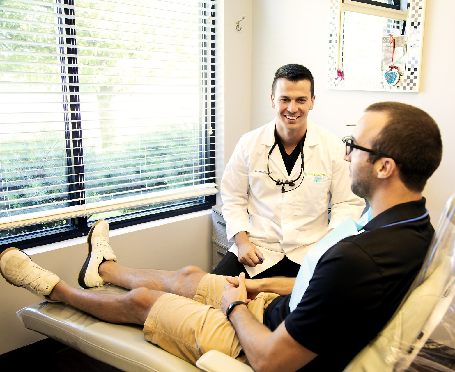 A healthcare professional in a white coat smiles at a patient sitting in a clinical exam chair.