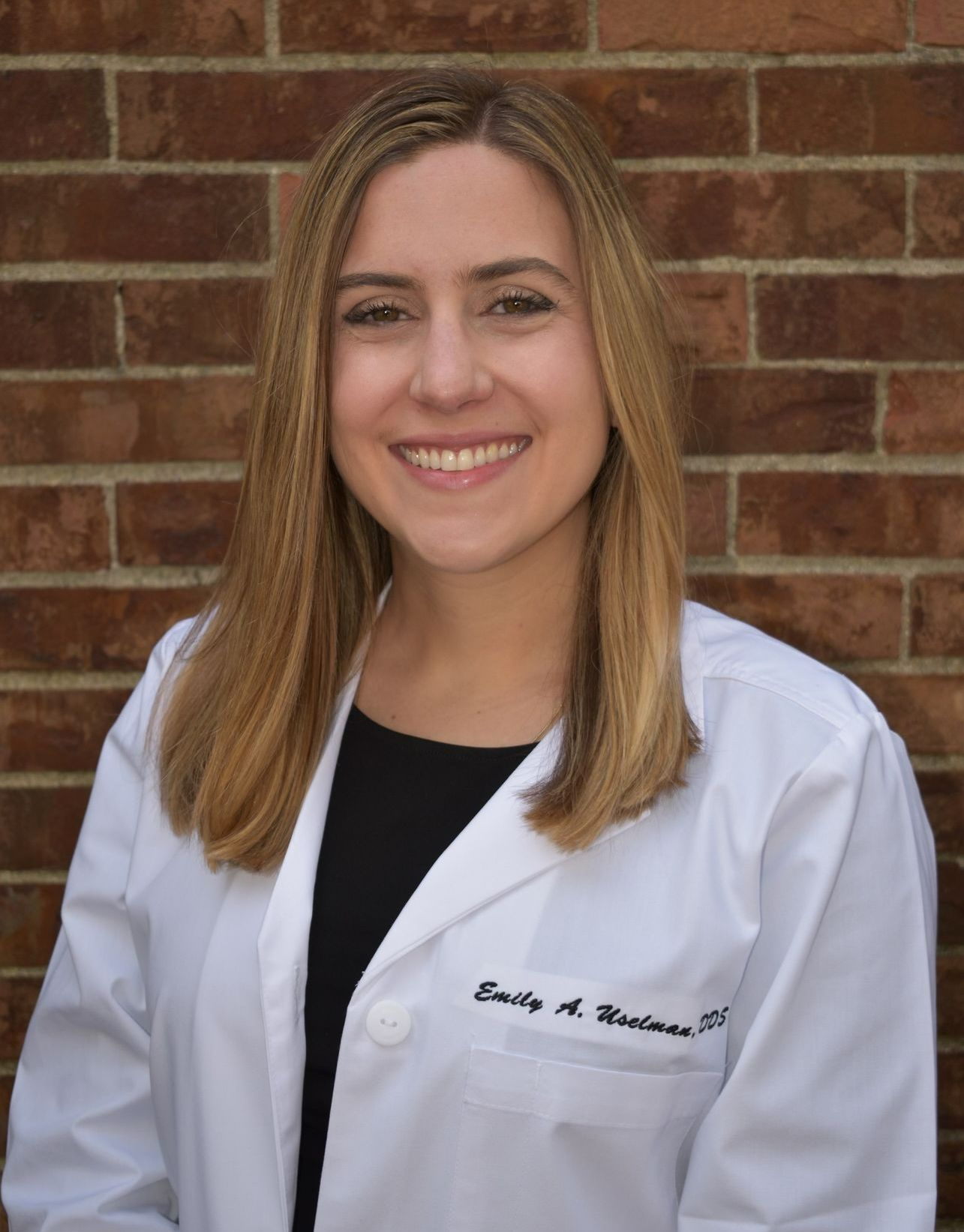 A smiling professional wearing a white lab coat with a name tag, standing in front of a brick wall.