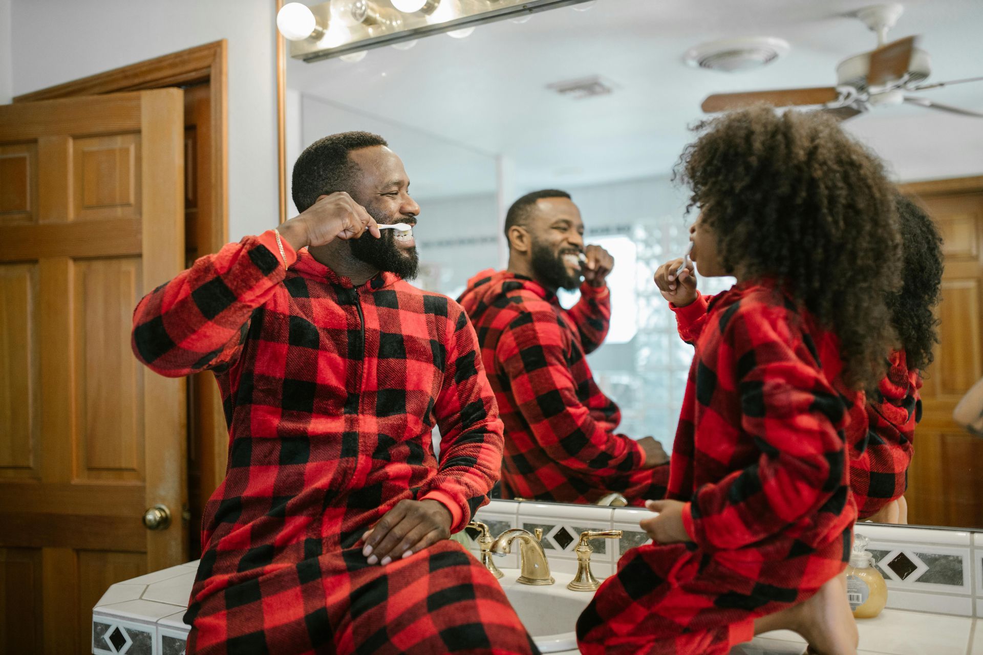 A parent and child in matching red and black plaid pajamas brush their teeth together in a bathroom.