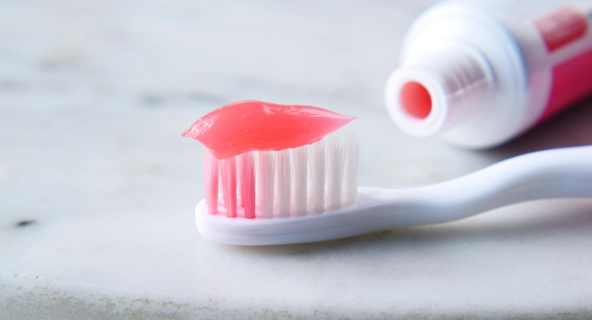 A white toothbrush with pink toothpaste sitting on its bristles next to an open tube of pink toothpaste on a marble surface.