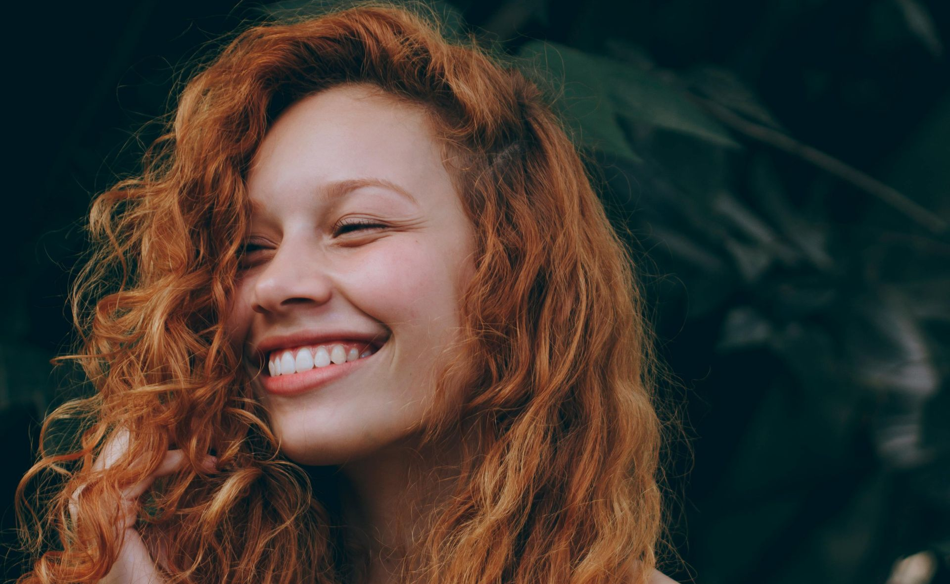 Smiling person with long, curly reddish-brown hair, set against a dark, out-of-focus background of foliage.