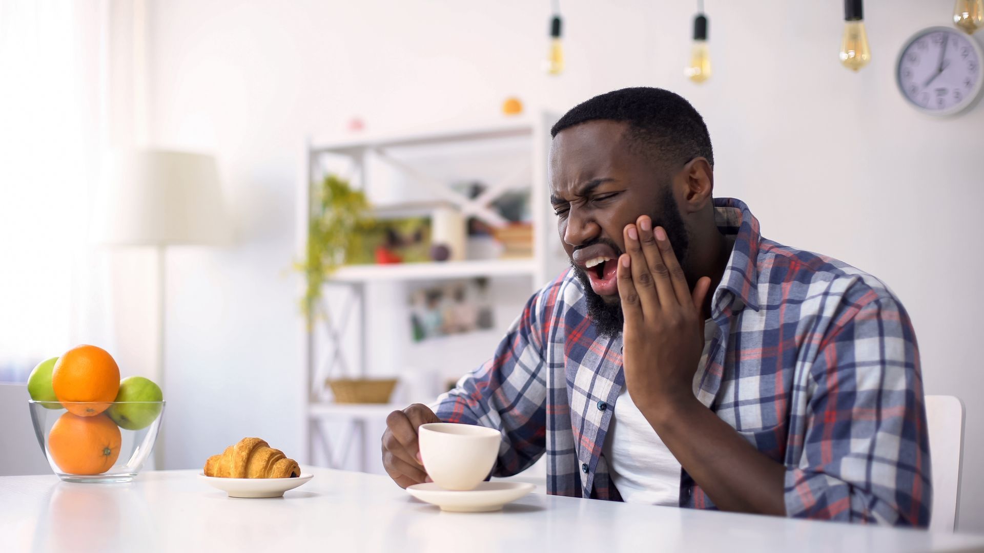 A person sitting at a table with fruit and a croissant, reacting in pain while holding their jaw after sipping a drink.
