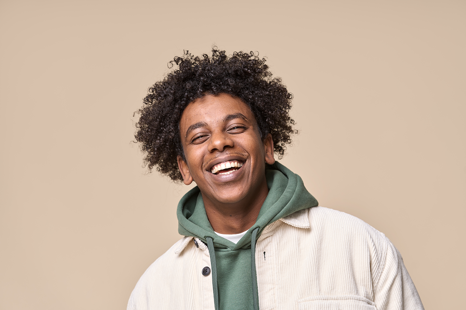 A person with curly hair smiling broadly, wearing a green hooded sweatshirt under a light-colored jacket against a tan wall.