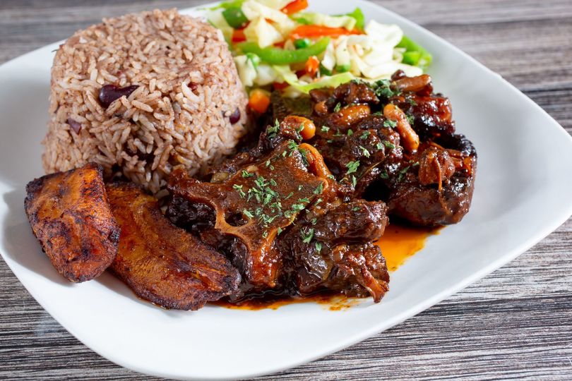 Plate of oxtail stew with rice and peas, fried plantains, and sauteed vegetables.